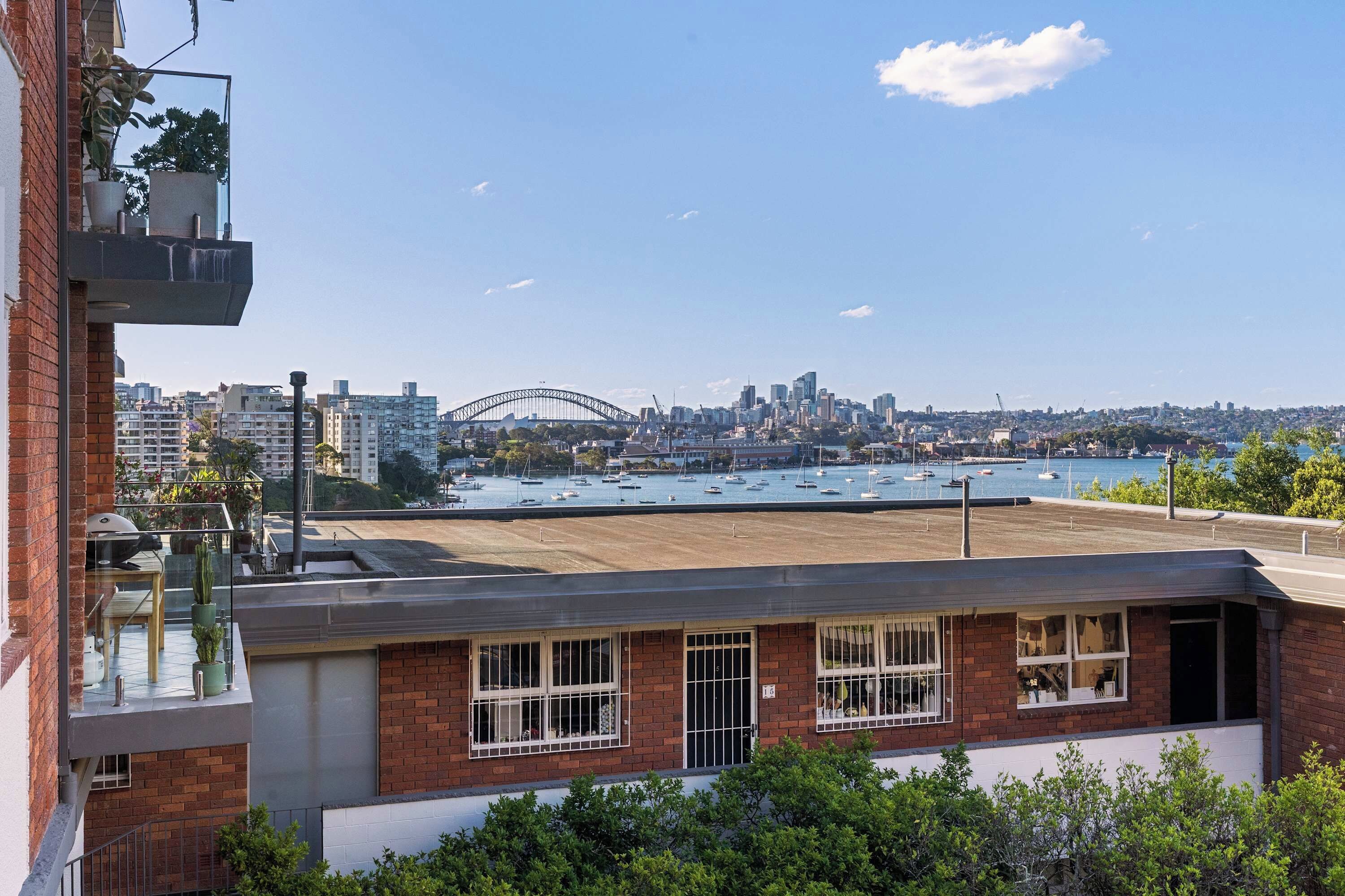 A building with a rooftop terrace overlooking a harbor with boats and a city skyline, including a prominent bridge.