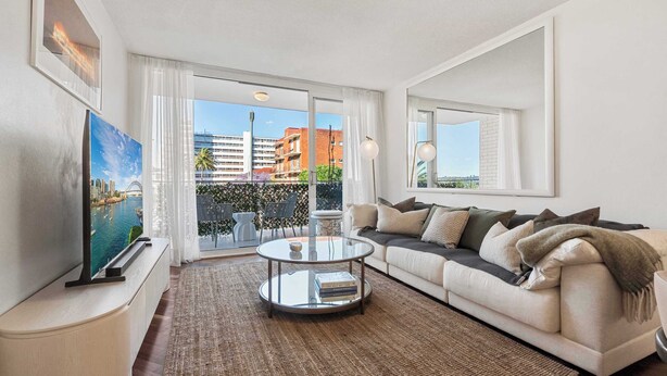 A modern living room with a sofa, a coffee table, and a TV on a stand. There is a large window with a view of buildings and a balcony. - Elegance in Darling Point (Darling Point)
