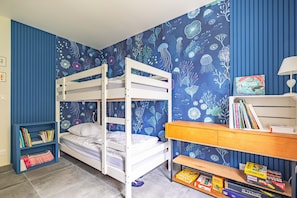 Een slaapkamer met stapelbed, blauw bloemenbehang, een boekenkast en een bureau met boeken en een lamp.