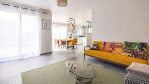 Una sala de estar moderna con una mesa de centro de vidrio, un sofá amarillo con cojines estampados, y un comedor al fondo.