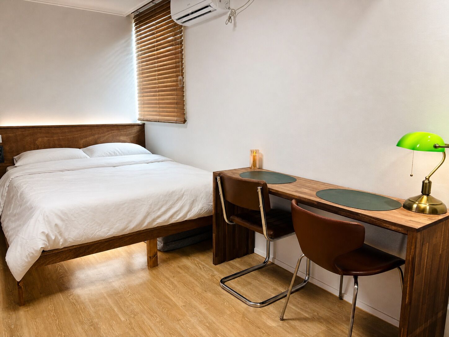 A bedroom with a bed, a desk with two chairs, a lamp, and an air conditioner.
