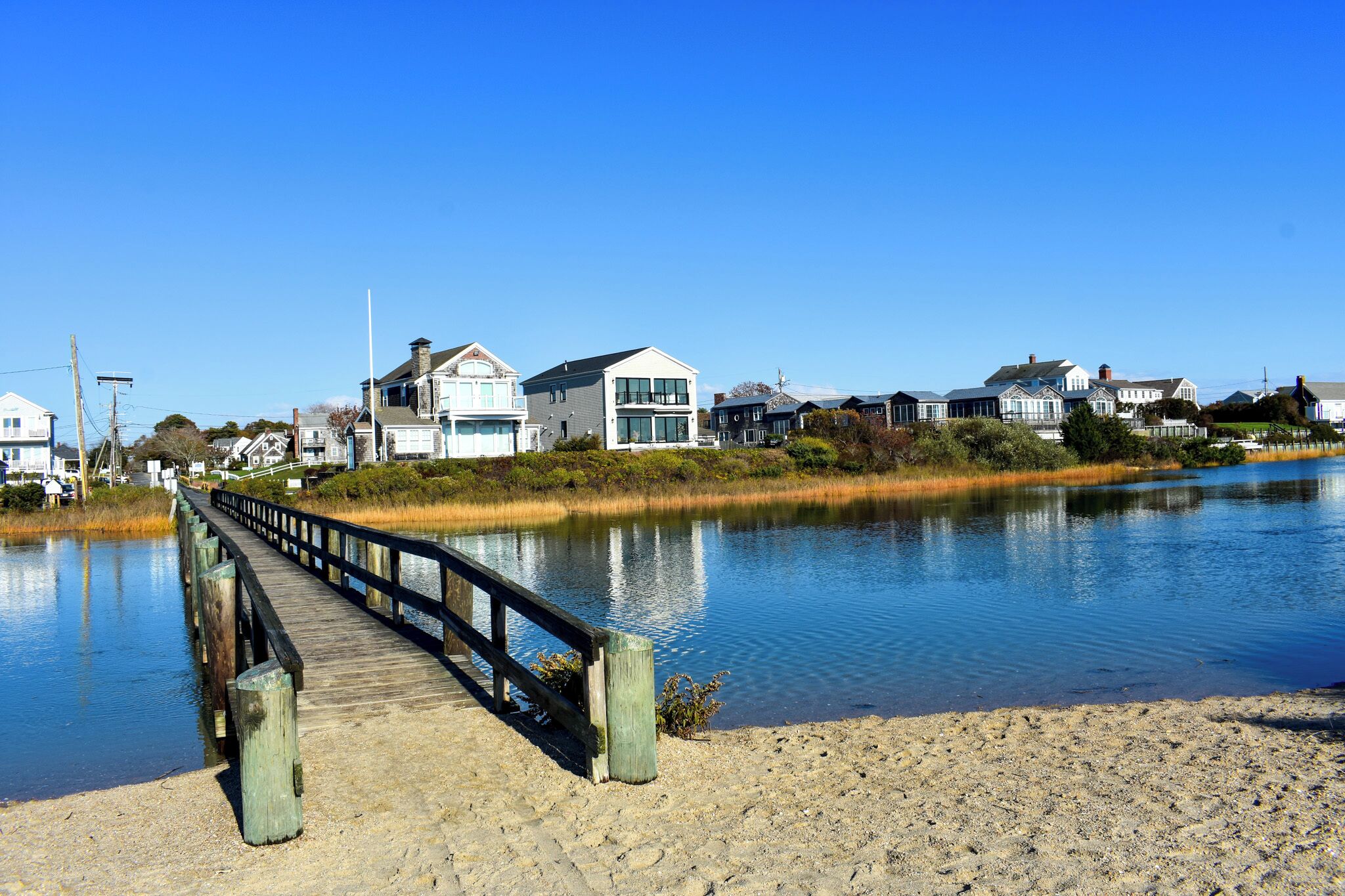 A wooden pier leading to a sandy beach with houses in the background.