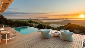 A wooden deck with a pool, two chairs, and a table overlooking a beach at sunset. - Mbizi Lagoon House (Hermanus)