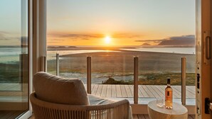 A balcony with a view of the sunset over the sea, a chair, and a bottle of wine on a small table. - Mbizi Lagoon House (Hermanus)