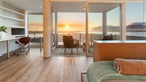 A modern hotel room with a wooden floor, a desk with a chair, a bed with a rolled towel, and a balcony with a view of the sunset over the water. - Mbizi Lagoon House (Hermanus)