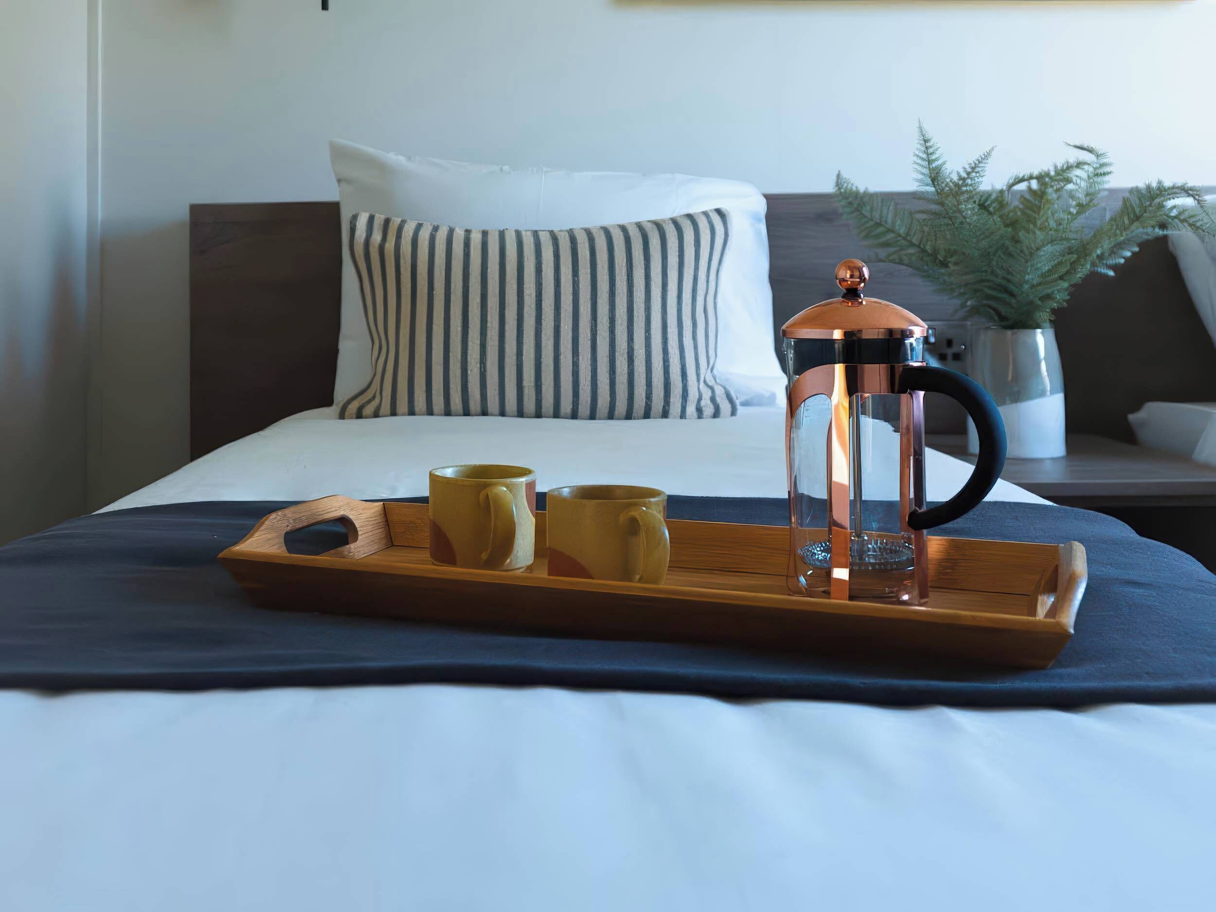 A neatly made bed with a tray holding a coffee maker, two mugs, and a potted plant.