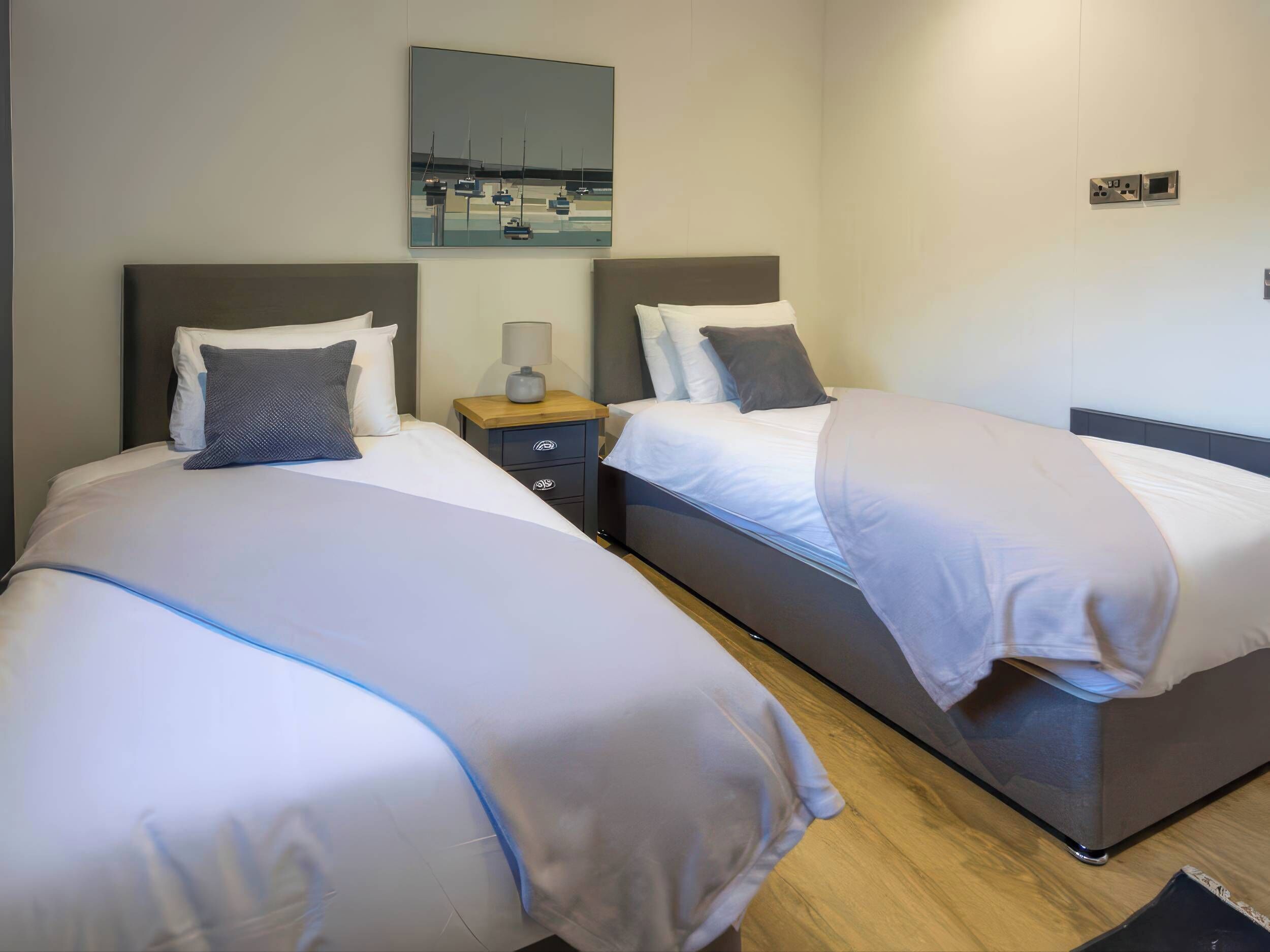 A hotel room with two beds, a nightstand with a lamp, a wall-mounted TV, and a painting of boats.