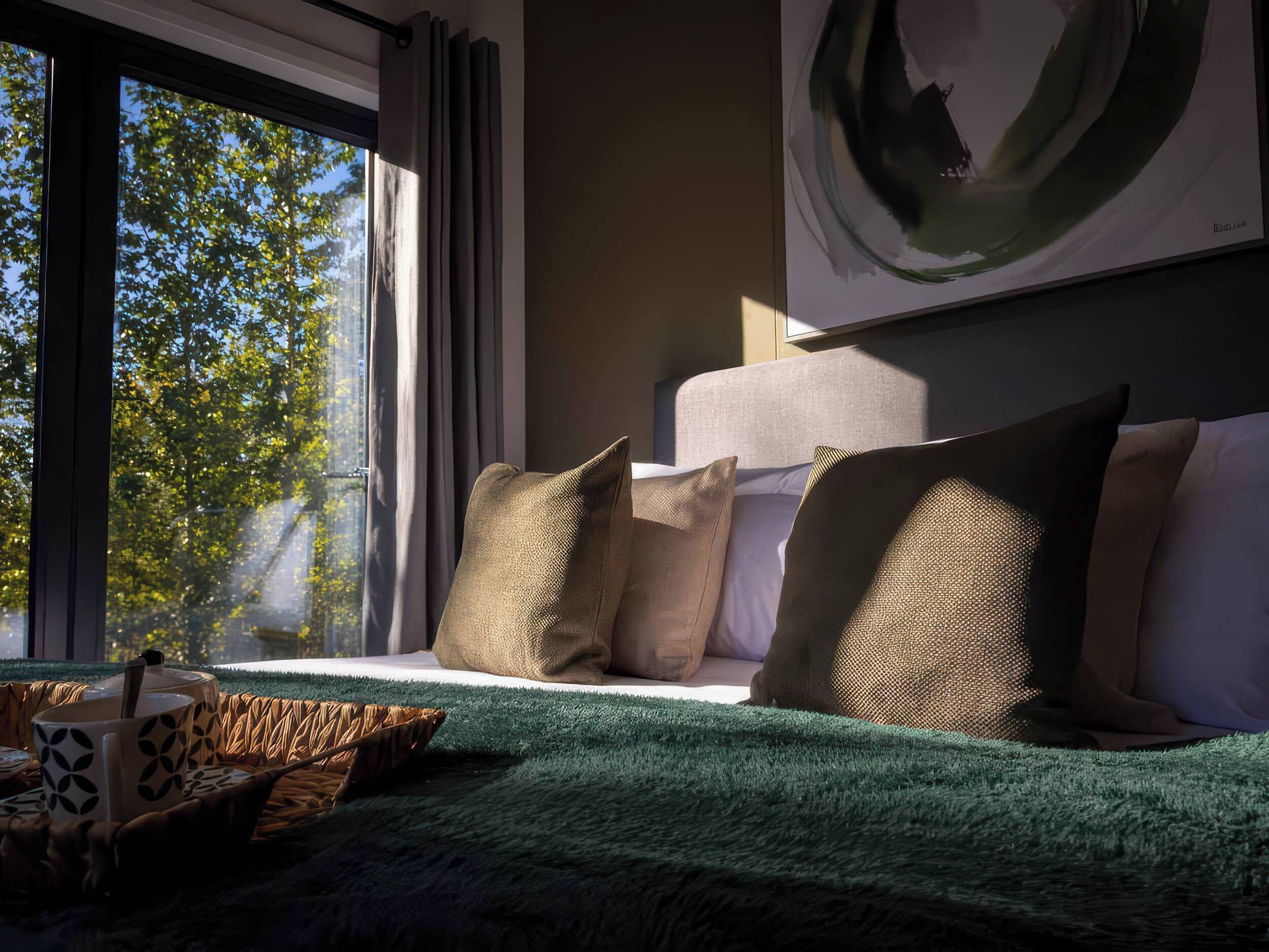 A modern bedroom with a large window, a bed with green bedding, and a view of trees outside.