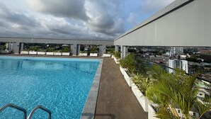 A rooftop pool with clear blue water, surrounded by potted plants and overlooking a cityscape.