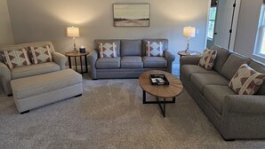 A living room with a grey sofa set, two side tables with lamps, a coffee table, and a painting on the wall. - New Home with Game Room and Nature Trails in Port Huron (Port Huron)