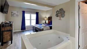 Un dormitorio con una bañera grande, una cama con cubrecama azul y blanco, un aparador de madera, una ventana con cortinas azules y un televisor montado en la pared.