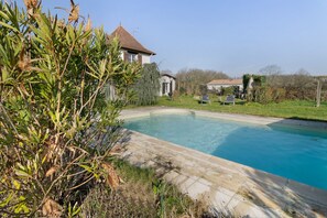 Une piscine avec une terrasse carrelée, entourée de verdure et une maison avec un toit en tuiles.