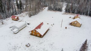 Un paysage enneigé avec plusieurs bâtiments en bois, dont une grande grange et de plus petites maisons, le tout entouré d’une forêt dense.