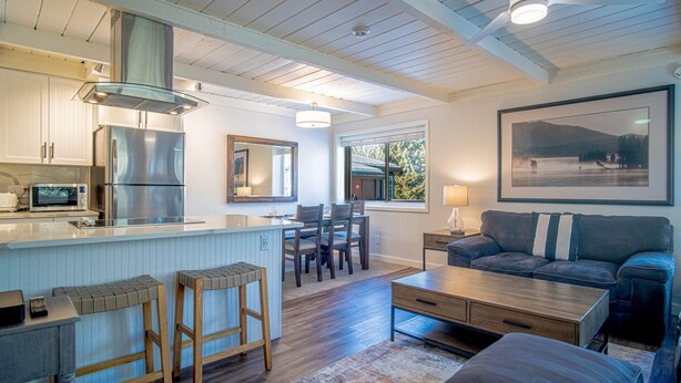 A modern kitchen with a stainless steel refrigerator, wooden bar stools, and a dining area with a table and chairs. - Atelier Allure - 2BD Mountain View + Free Resort Amenities (Sun Valley)