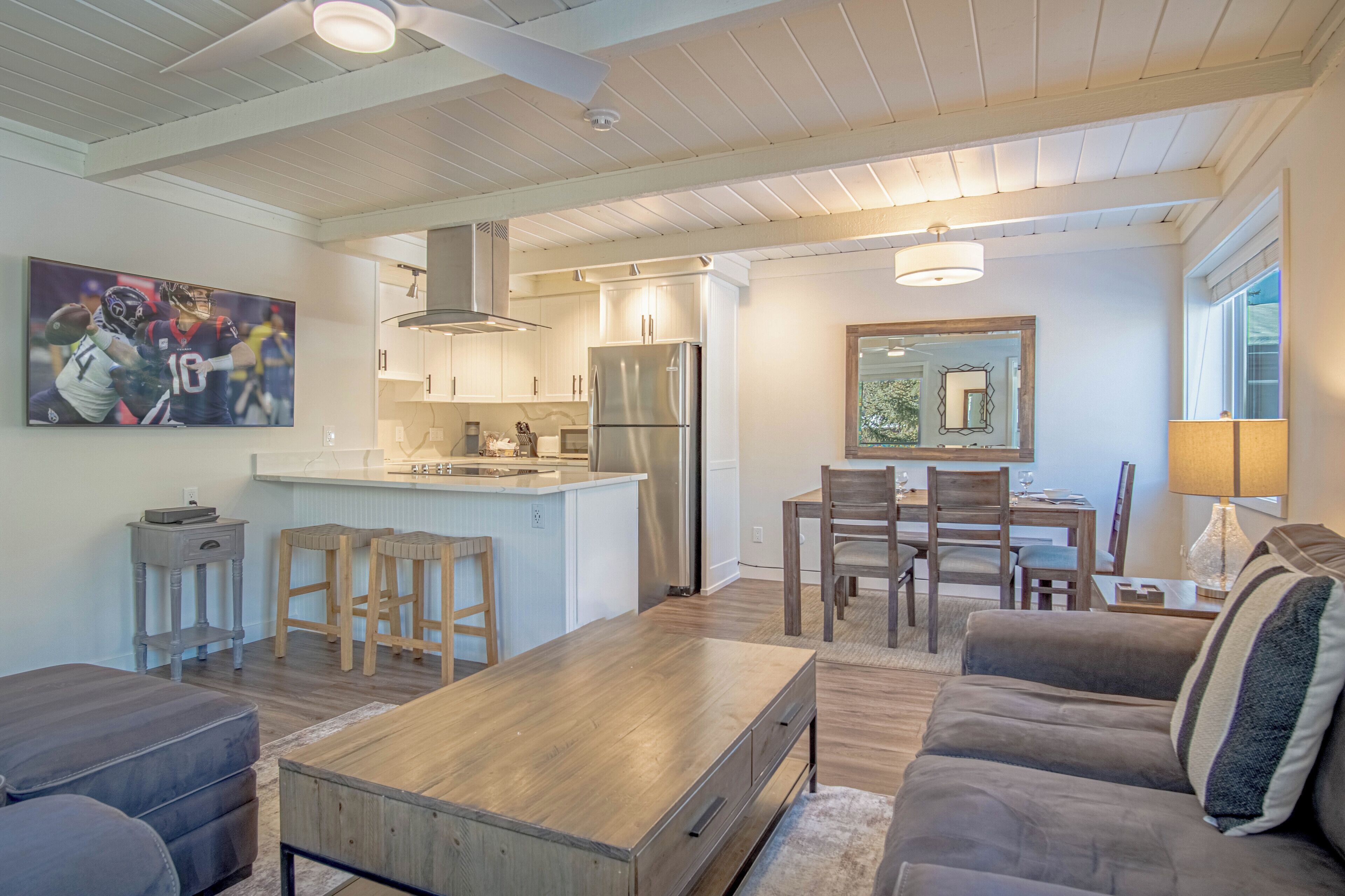 Una cocina moderna con una isla central, taburetes de madera, un comedor con mesa y sillas, y una sala de estar con sofá y mesa de centro.