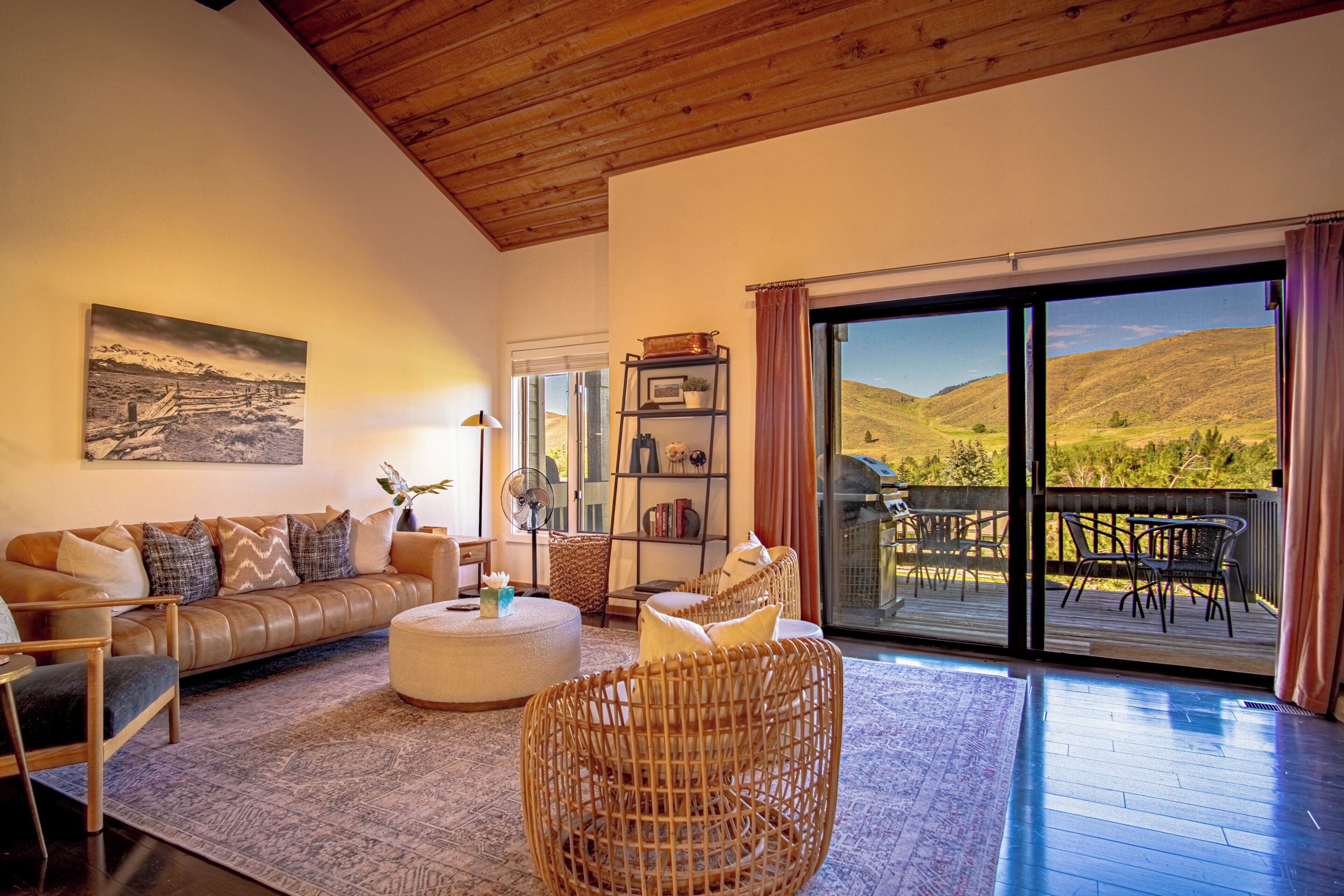 A spacious living room with a large window, a sofa, a wicker chair, and a view of a mountainous landscape.