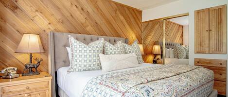 A bedroom with a wooden slat ceiling, a tufted headboard, multiple pillows, a patterned bedspread, a bedside lamp, a telephone, and a wooden cabinet.