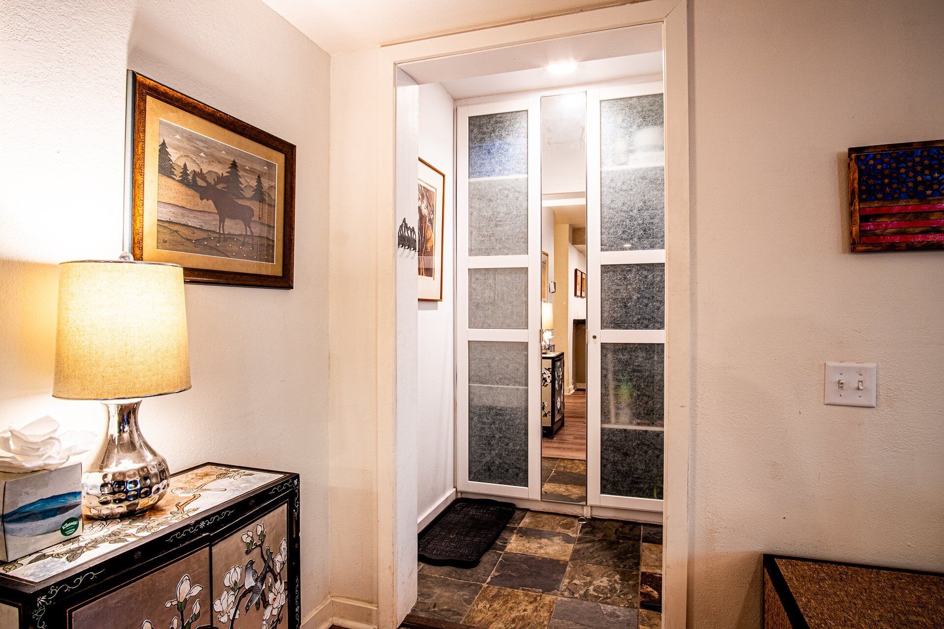 A hallway with a lamp, framed artwork, and a door leading to another room.