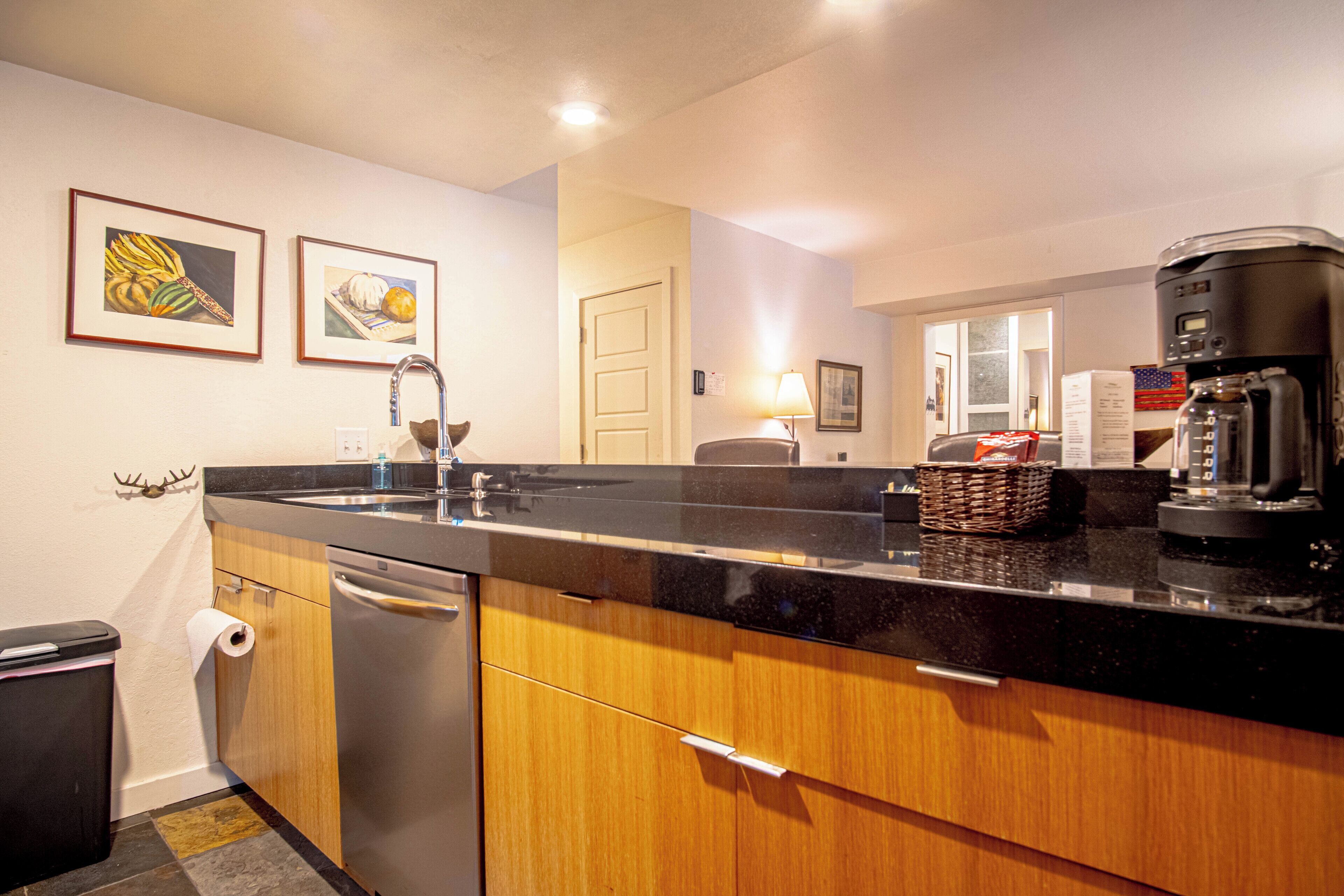 A modern kitchen with wooden cabinets, a stainless steel dishwasher, a black countertop, and a coffee maker.
