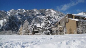 A snow-covered landscape with a building featuring a modern design, surrounded by snow-laden trees and mountains in the background. - Sv139 - Walk to Lifts & Town - Hot Tub & Pool (Ketchum)