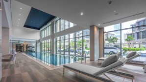 A modern indoor swimming pool area with lounge chairs and a view of the city through large windows. - StayShort - Canal View 2BR in the Heart of Business Bay - Sleeps 6! (Business Bay)