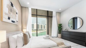 A modern bedroom with a large bed, a nightstand, a dresser, a mirror, and a view of a building through the window. - StayShort - Canal View 2BR in the Heart of Business Bay - Sleeps 6! (Business Bay)