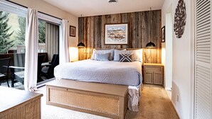 Ein Schlafzimmer mit einem großen Bett, Holzwandverkleidung, einem Bild über dem Bett, zwei Nachttischlampen und Blick ins Freie durch eine Schiebetür.