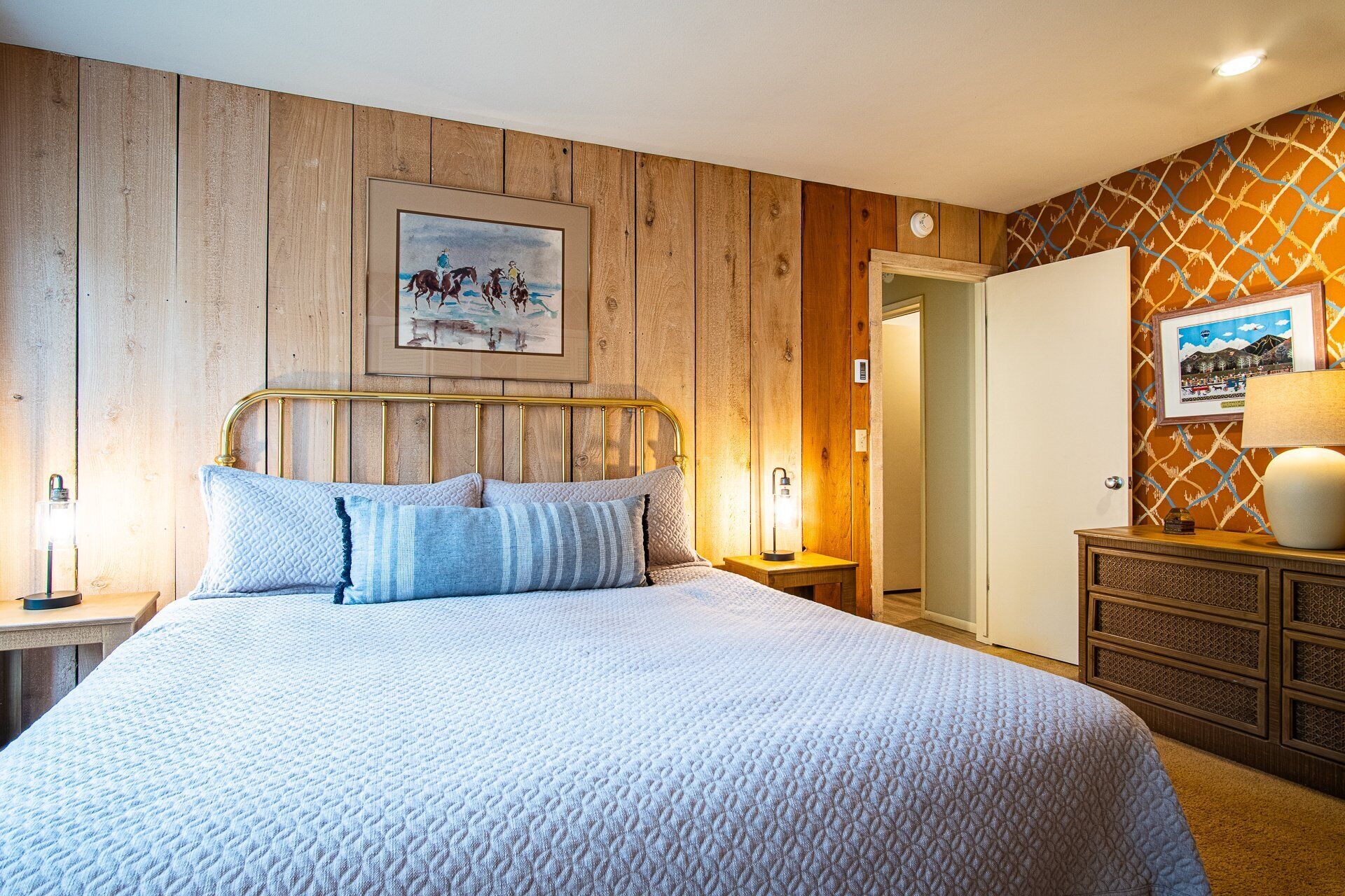 Une chambre avec un mur lambrissé en bois, un lit recouvert d’une couette blanche, deux lampes de chevet, un tableau encadré au-dessus du lit, une armoire et une porte donnant sur une autre pièce.