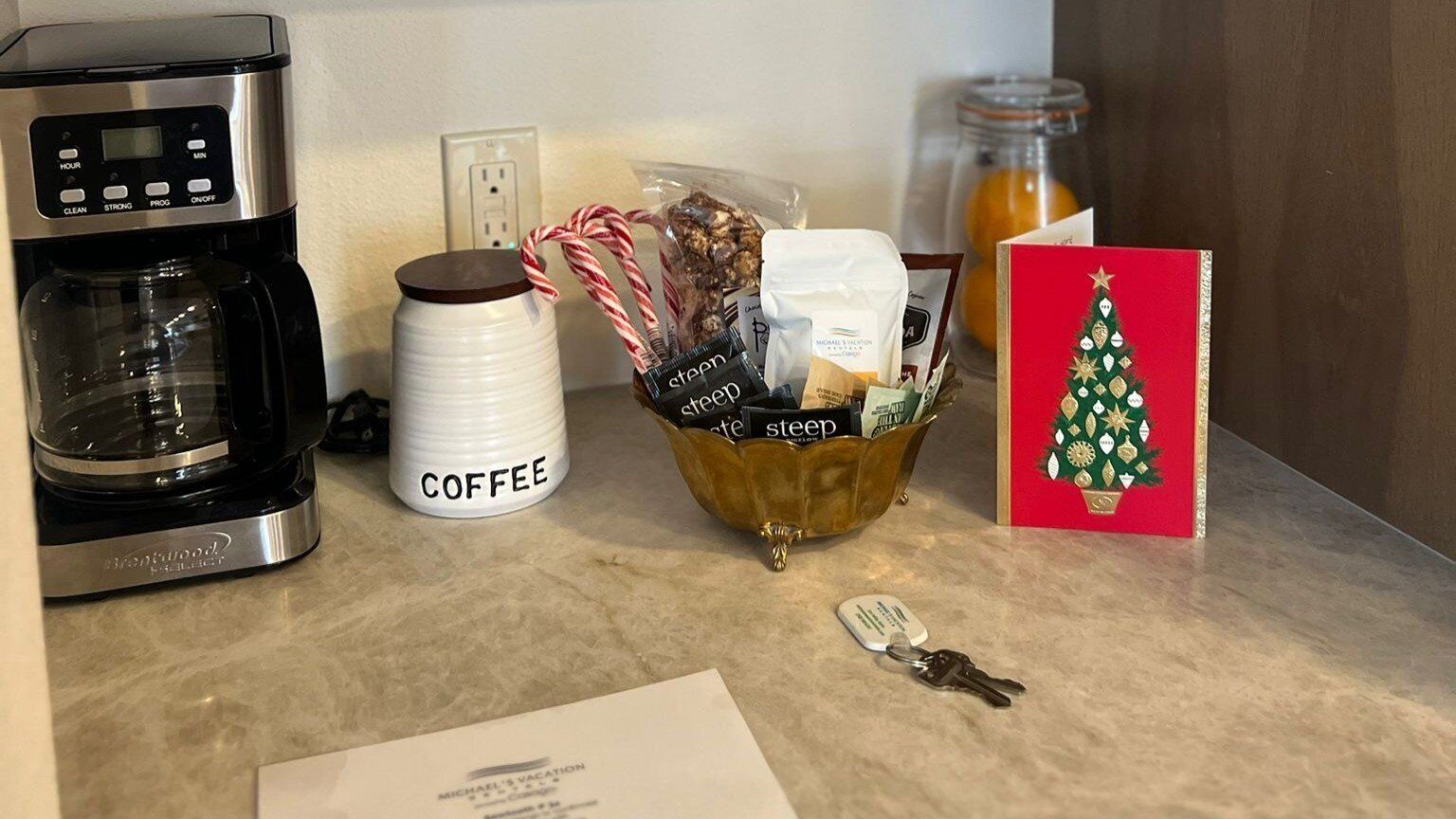 A kitchen counter with a coffee maker, a coffee container, a basket of snacks, a Christmas card, and a keychain.