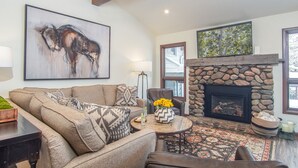A living room with a stone fireplace, a large abstract painting, and a cozy seating area. - Skiway Chalet - Next to Warm Springs Lifts - Close to Natural Hot Springs (Ketchum)