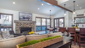 A spacious living room with a stone fireplace, a large sofa, and a dining area with a wooden table and chairs. - Skiway Chalet - Next to Warm Springs Lifts - Close to Natural Hot Springs (Ketchum)