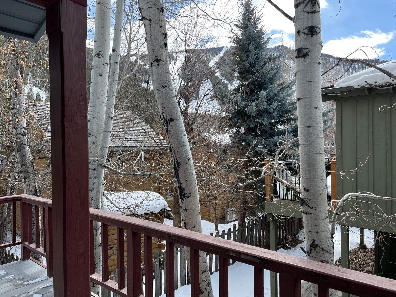 A wooden deck with a railing, surrounded by trees and a building with a sloped roof.