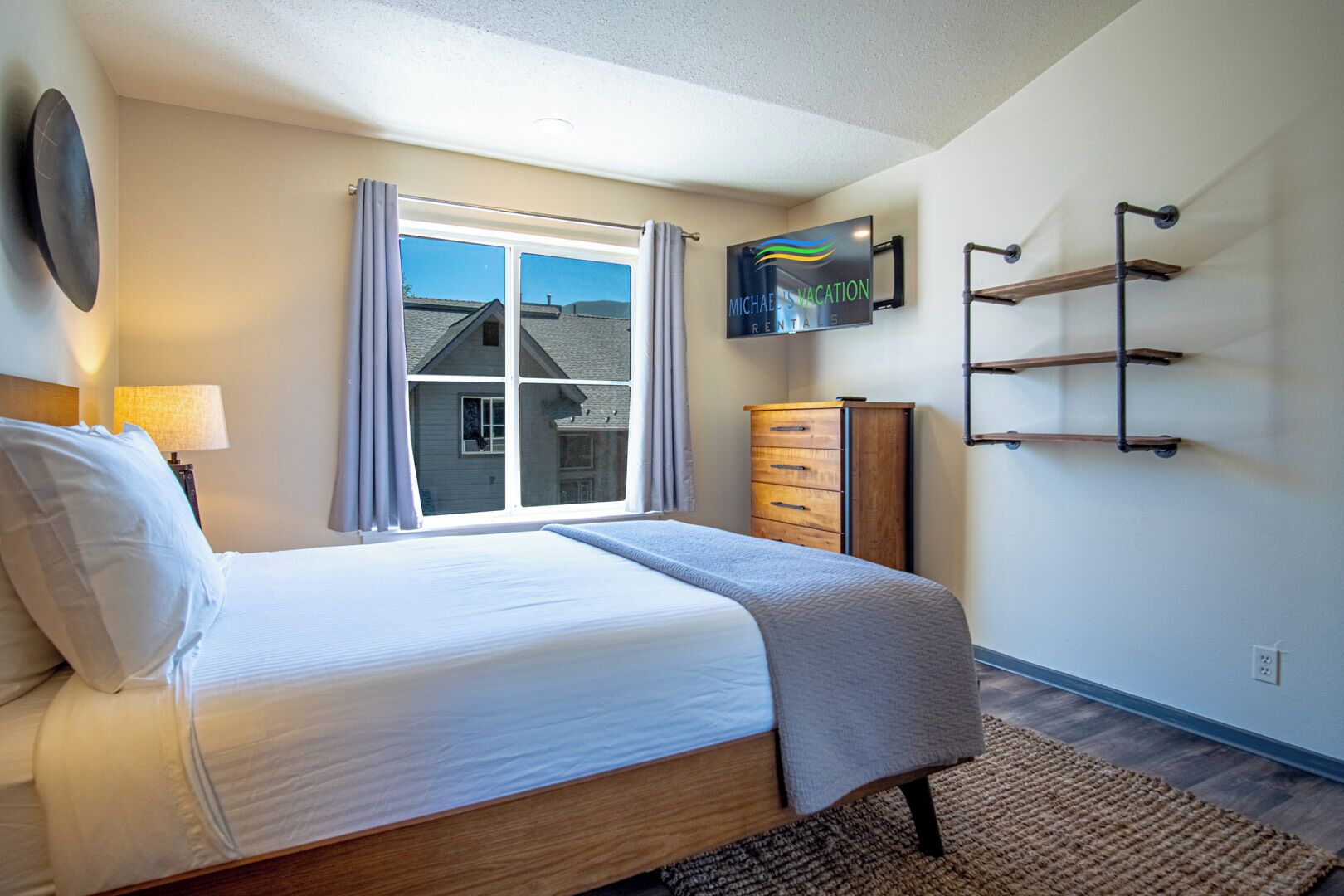 A bedroom with a bed, a dresser, a TV mounted on the wall, and a window with curtains.