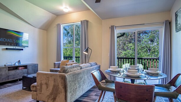 A modern living room with a grey sofa, a dining table set for four, and large windows offering a view of greenery. - Della View 2BR Condo - In the Heart of Historic Downtown Hailey! (Hailey)