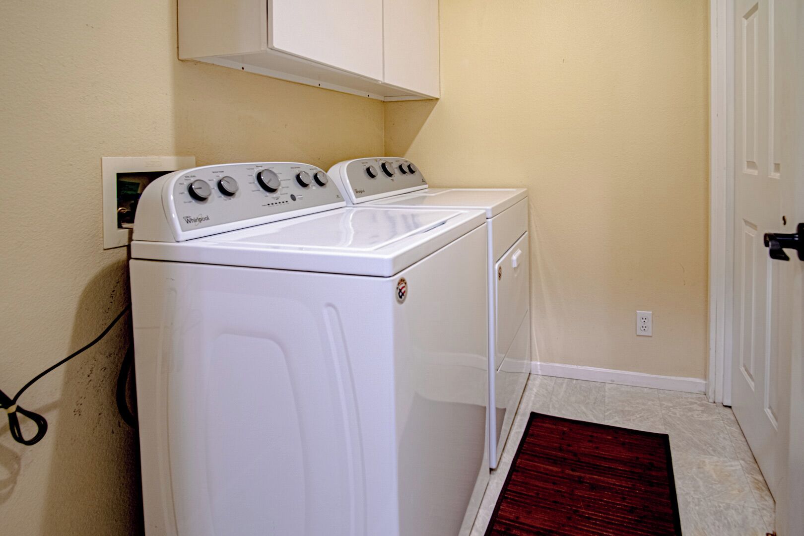 A compact laundry room with a front-loading washer and dryer, a white cabinet above the washer, and a red doormat at the entrance.