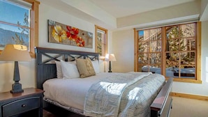 A bedroom with a large bed, two bedside tables, a window with a view of trees, and a painting on the wall. - Elkhorn Springs Sanctuary - Pool/hot Tub/underground Parking (Sun Valley)