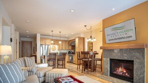 A modern living room with a fireplace, striped sofa, and a dining area with wooden chairs. - Elkhorn Springs Sanctuary - Pool/hot Tub/underground Parking (Sun Valley)