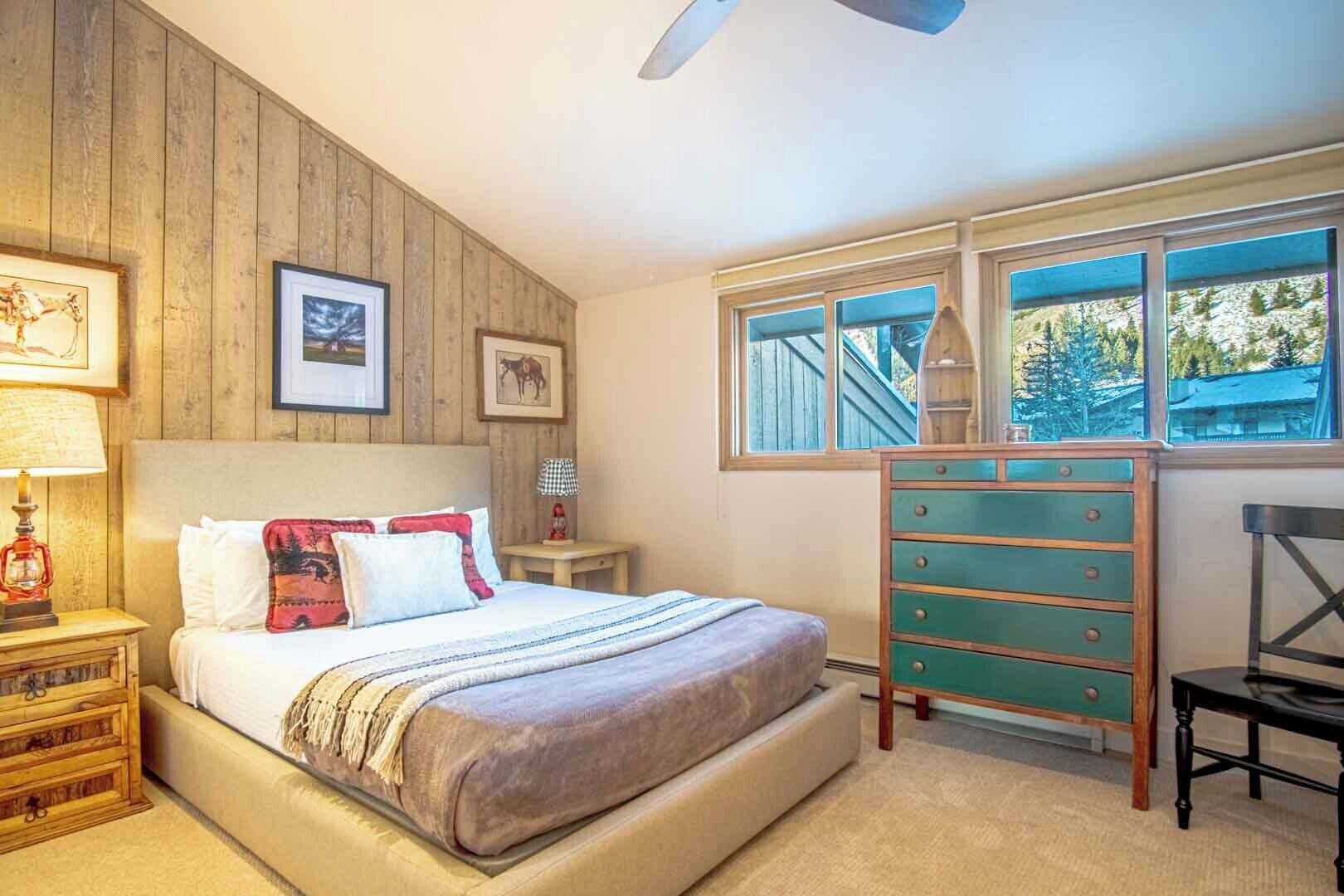 Un dormitorio con pared de madera, una cama con muchos almohadones, un aparador, una silla y una ventana que ofrece vistas a árboles y un edificio.