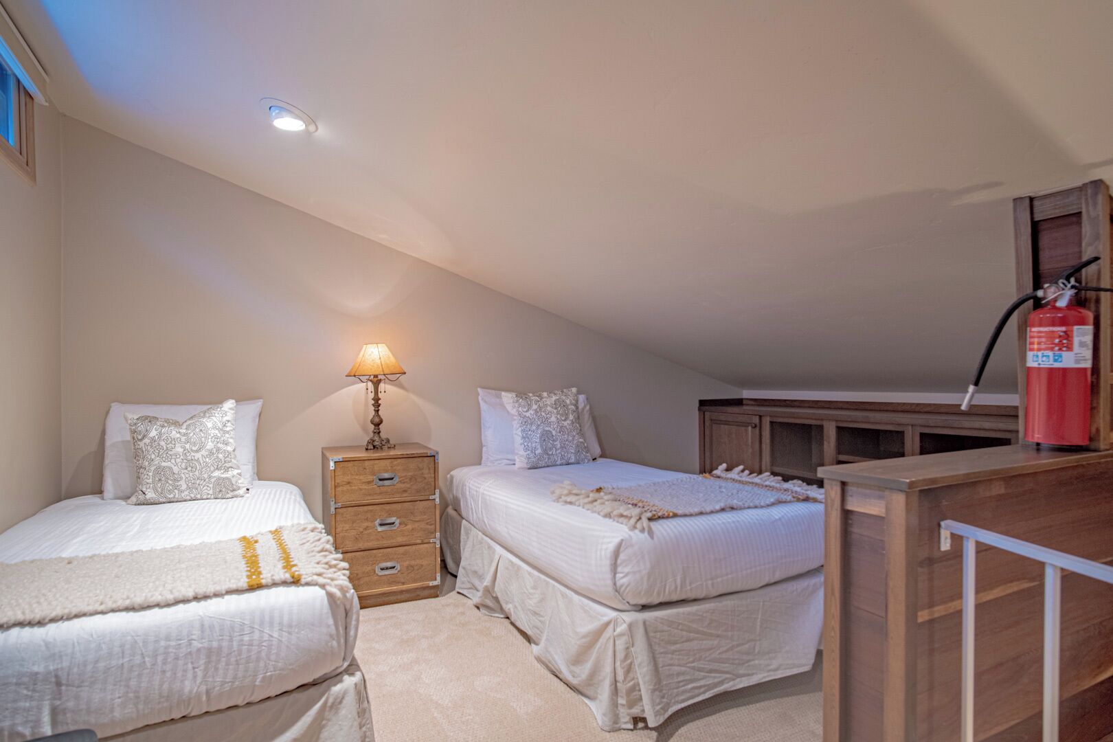 A bedroom with two beds, a nightstand with a lamp, a wooden dresser, and a fire extinguisher.