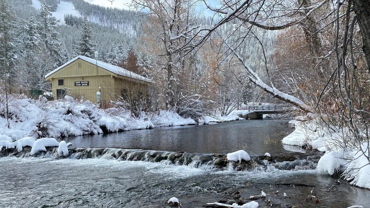 A snow-covered landscape with a wooden building labeled "Snow Making Pump House" and a bridge over a river.