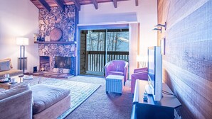 A cozy living room with a stone fireplace, wooden beams, a television, and a comfortable seating area. - Snow Creek 1553 - Ski-in/ski-out + Pool & Hot Tub (Sun Valley)