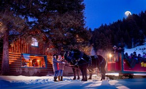 A wooden cabin with a lit interior, a sleigh with passengers, and a horse. - Snow Creek 1553 - Ski-in/ski-out + Pool & Hot Tub (Sun Valley)