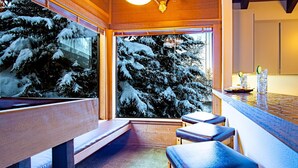 A cozy cabin interior with a wooden bench and a bar area, featuring a large window with a snowy forest view. - Snow Creek 1553 - Ski-in/ski-out + Pool & Hot Tub (Sun Valley)