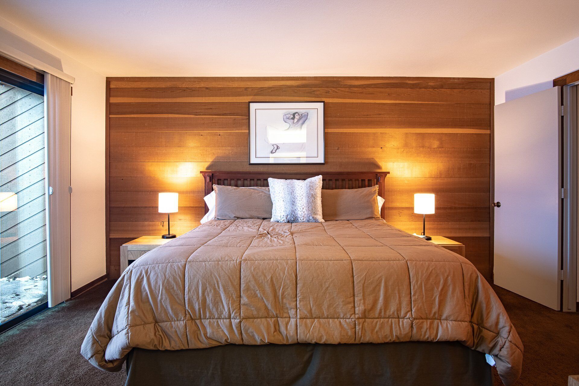 A bedroom with a wooden headboard, a framed picture, two bedside lamps, and a bed with a beige duvet.