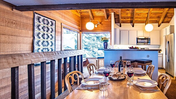 A wooden dining area with a table set for four, a kitchen in the background, and a view of the outdoors. - Snow Creek 1553 - Ski-in/ski-out + Pool & Hot Tub (Sun Valley)