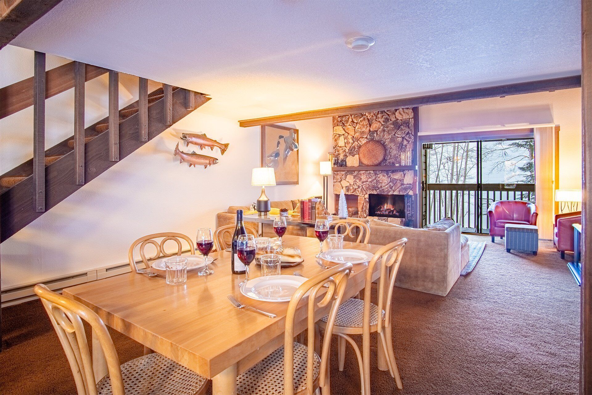 A dining area with a wooden table set for four, surrounded by wicker chairs. A stone fireplace and a large window with a view of trees are visible in the background.