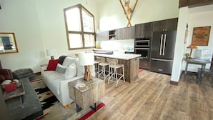 A modern living room with a sofa, coffee table, and a dining area with bar stools. A kitchen with dark cabinets and stainless steel appliances is adjacent. - Snow Creek 1553 - Ski-in/ski-out + Pool & Hot Tub (Sun Valley)