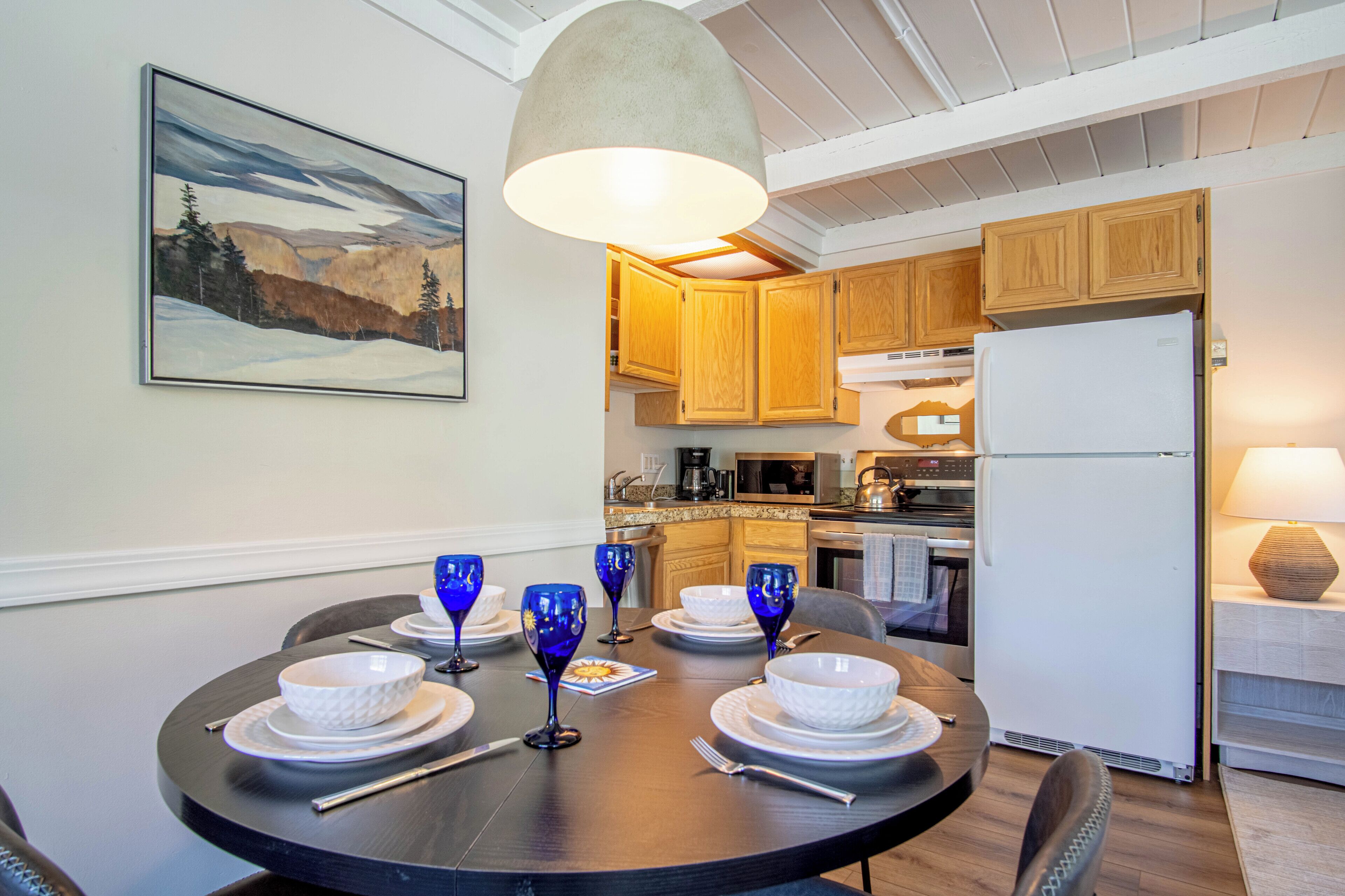 A dining area with a round table set for four, wooden cabinets, a white refrigerator, and a painting on the wall.