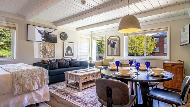 A cozy living room with a bed, sofa, coffee table, and dining table with chairs. There are framed pictures on the walls, a clock, and a window with a view of trees and buildings. - Atelier Escape - Sun Valley Studio + Resort Amenities (Sun Valley)