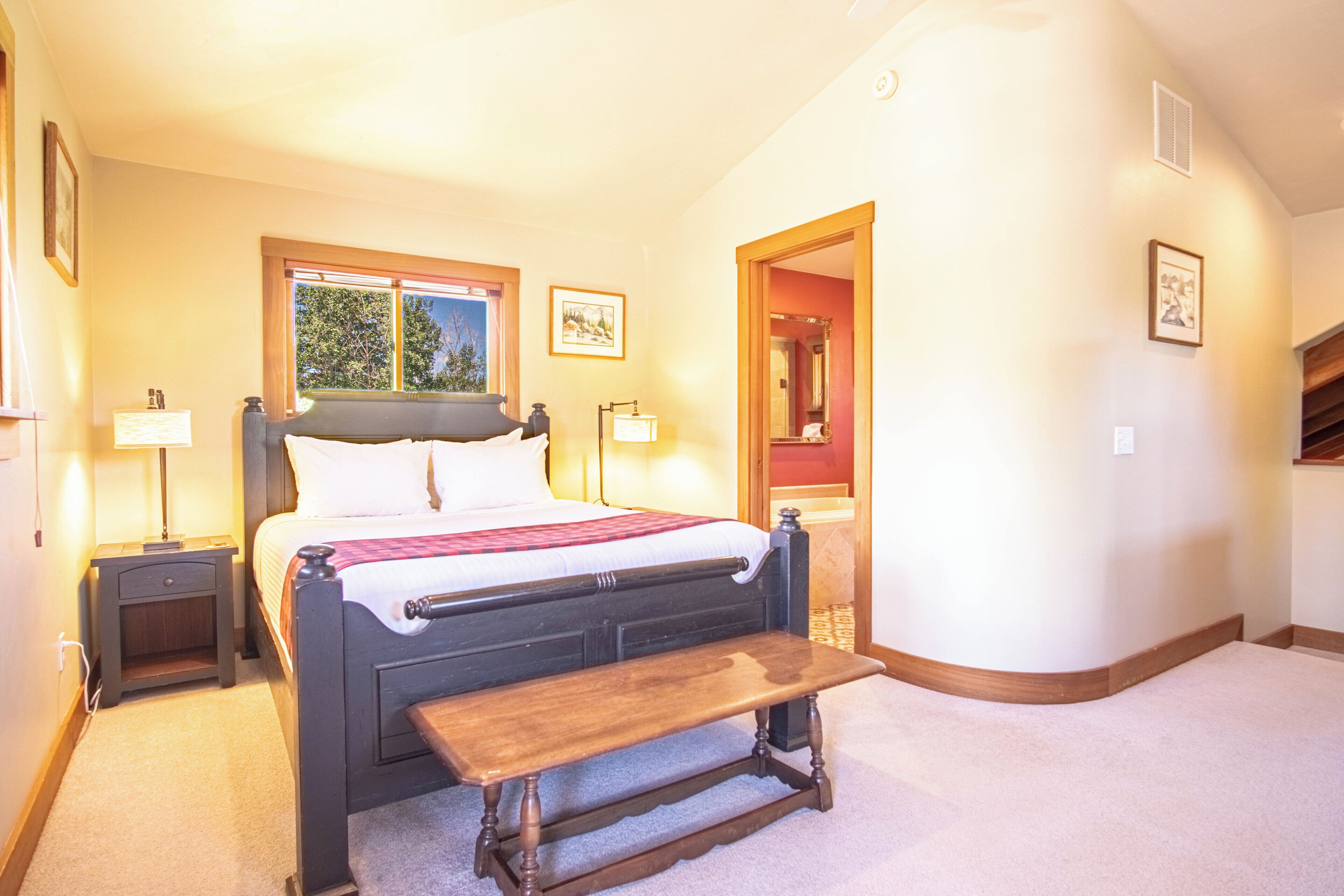 Une chambre à coucher comprenant un lit, des tables de chevet, un banc, une fenêtre et une porte donnant sur une salle de bain.